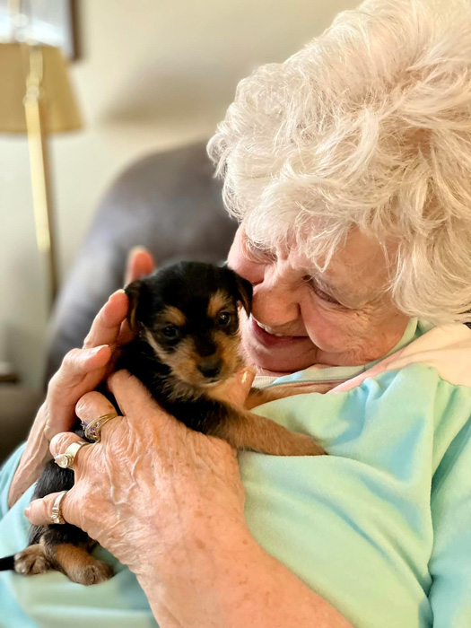 A senior resident with white hair smiles while holding a small puppy close to her chest, enjoying a heartwarming moment of companionship in a senior living community.