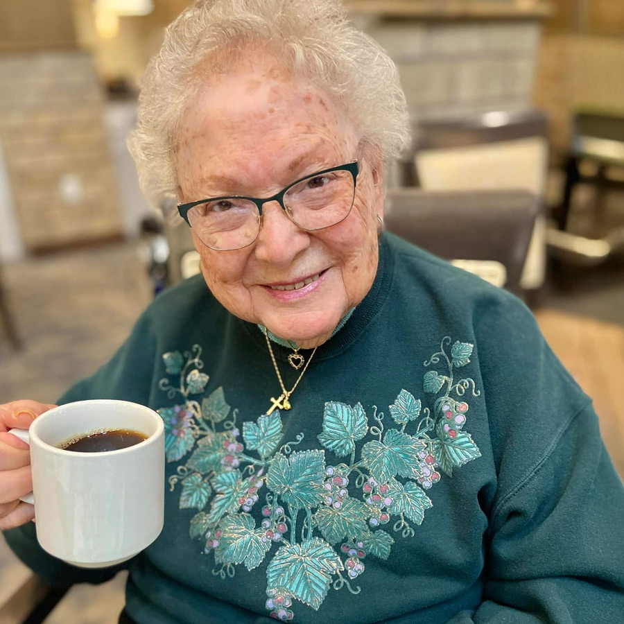 Senior resident smiling and holding a cup of coffee, wearing a green sweater adorned with floral embroidery and a gold necklace, enjoying a moment in a cozy indoor setting.