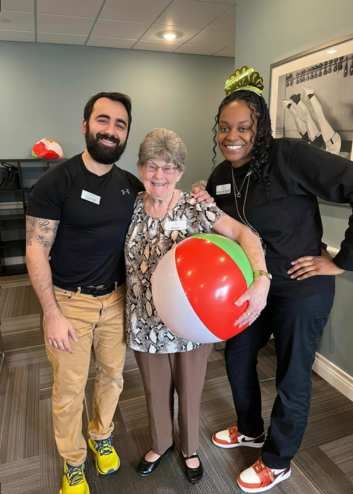 Smiling senior resident holding a beach ball stands between two staff members, one male and one female, during a festive indoor event, with all three posing happily together.