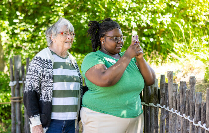 Two women enjoying a sunny day outdoors, with one taking a photo on her phone and the other smiling beside her, standing by a wooden fence surrounded by lush greenery.