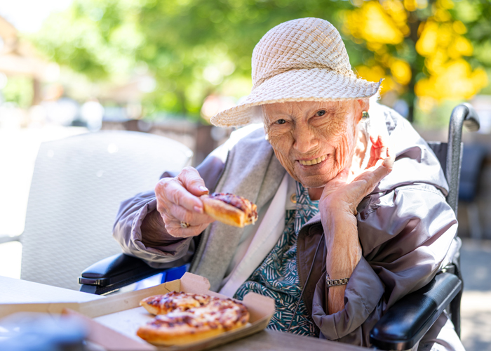 Senior resident in a sun hat smiles warmly while enjoying a slice of pizza outdoors, highlighting the joy of simple pleasures and vibrant living in a senior community.