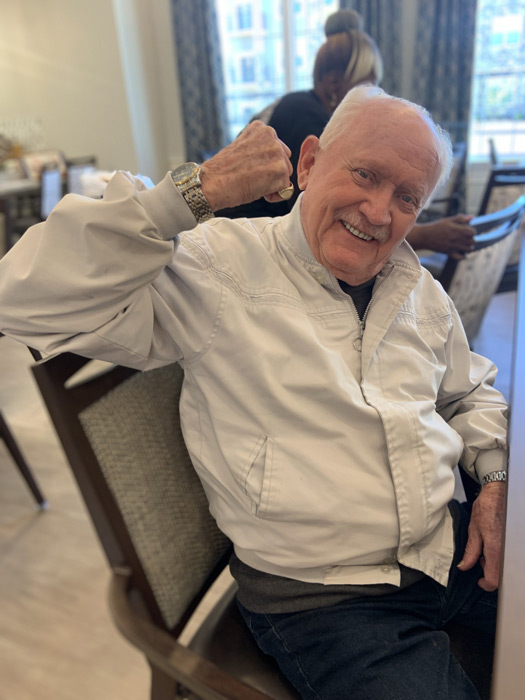 Smiling senior resident in a white jacket raises his arm in a playful flexing gesture while seated in a senior living dining area, exuding confidence and joy.