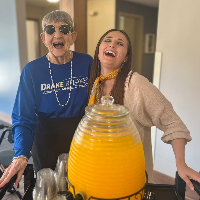 Two women, one a senior resident wearing sunglasses and pearls, share a joyful moment beside a large container of orange juice during a fun, casual gathering.