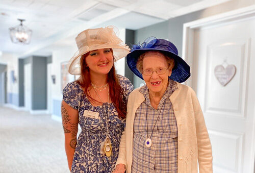 Senior resident and team member at a senior living community dressed in elegant outfits with hats, standing together in a well-lit hallway, sharing smiles during a special event.