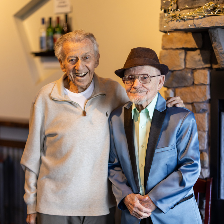 Two smiling senior residents standing together at a senior living community event, one in a light sweater and the other in a blue blazer and hat, enjoying a moment of friendship.