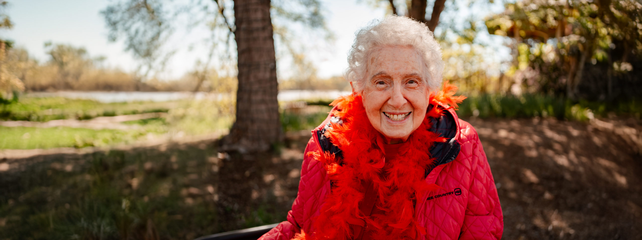 Smiling senior resident in a bright red jacket and feathered boa enjoying a sunny day outdoors, with trees and a serene lake in the background, exuding warmth and joy.