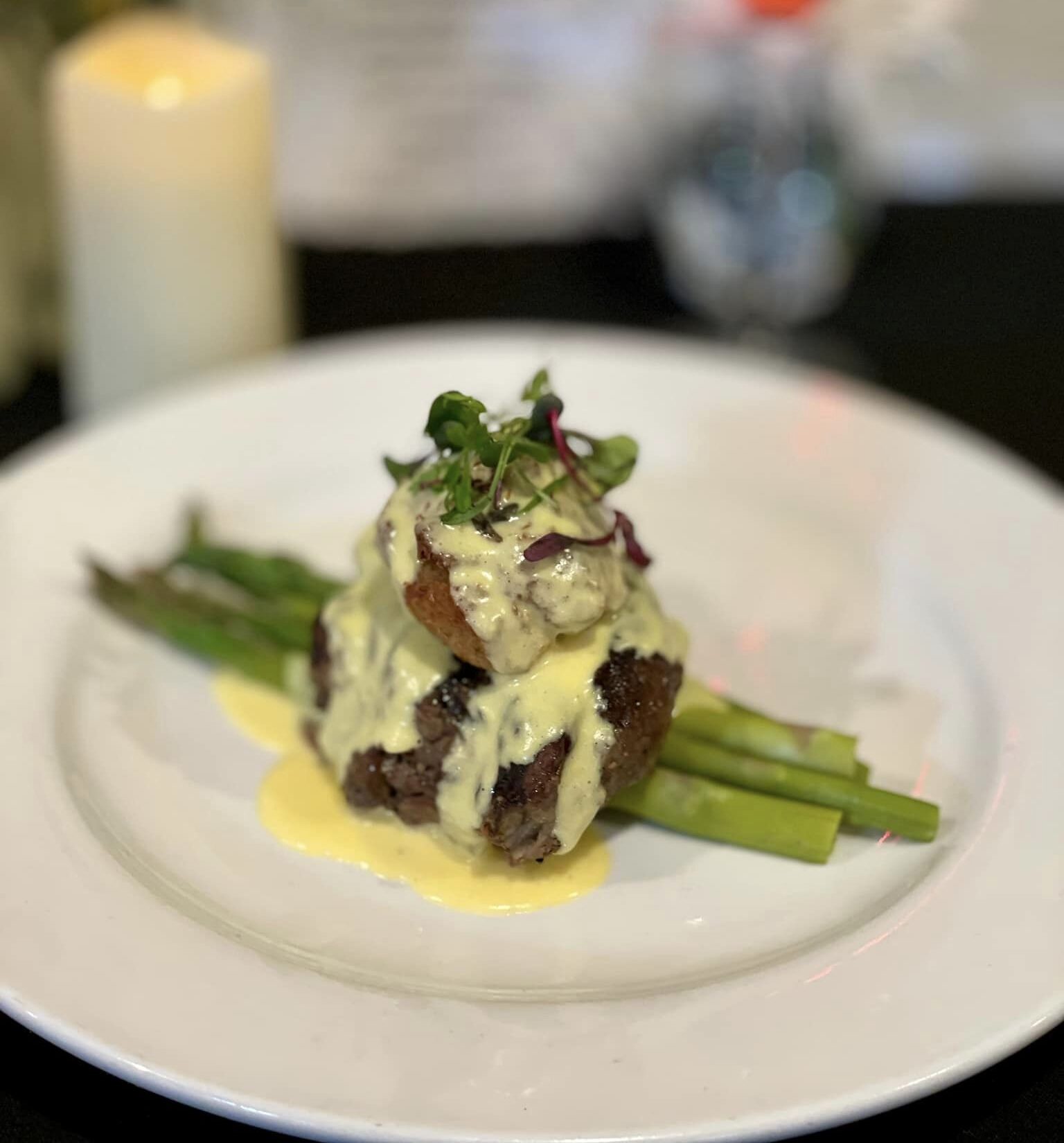 Plated gourmet steak with asparagus, topped with creamy hollandaise sauce and garnished with microgreens, set against a blurred background of a candle, flowers, and drink.