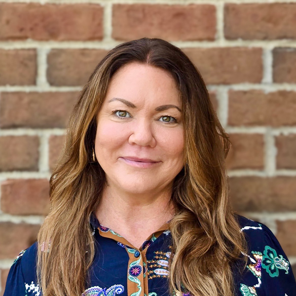 Nora Zands, Senior Living Director at Holliday Farms Senior Living, smiling in a navy paisley print blouse in front of a brick wall.