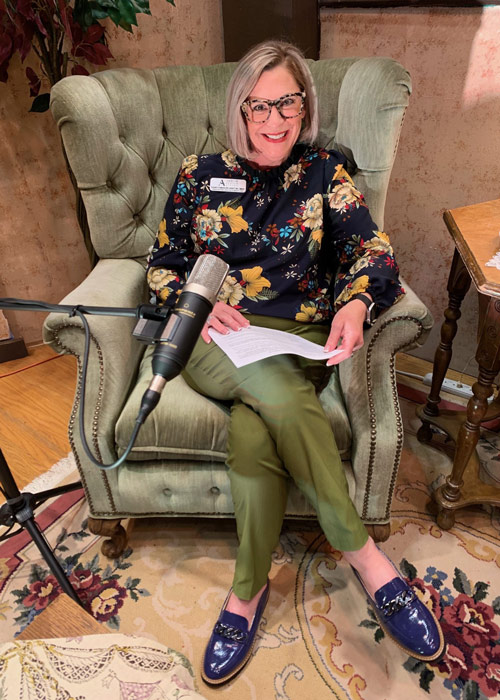 Elizabeth, one of the hosts of Thoughtful Connections: A Memory Care Podcast, sits comfortably in a green armchair with a microphone and notes in front of her during recording.