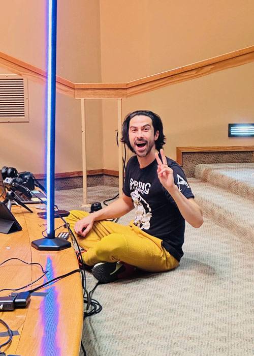 Producer Lenny Hernandez sits cross-legged on a carpeted stage, smiling and flashing a peace sign beside podcast lights during filming for Thoughtful Connections at Cottey College