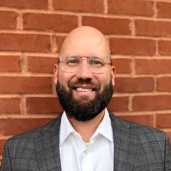 CJ Boswell, Executive Director at Holliday Farms Senior Living, smiling in a professional headshot, wearing glasses, a white shirt, and a gray blazer, standing in front of a red brick wall.