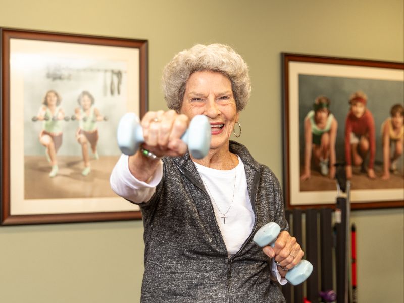 A senior woman holding small hand weights flexes one arm in front of herself in the community exercise room.