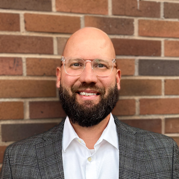 CJ Boswell, Executive Director at Holliday Farms Senior Living, smiling in a professional headshot, wearing glasses, a white shirt, and a gray blazer, standing in front of a red brick wall.