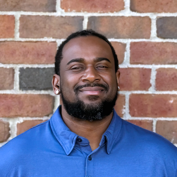 Daniel Thomas, Operations Coordinator at Holliday Farms Senior Living, smiling in a professional headshot, wearing a blue collared shirt, with a brick wall background.