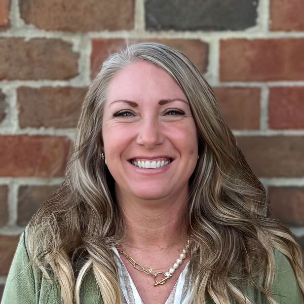Stephanie Garber, Senior Living Director at Holliday Farms Senior Living, smiling in a professional headshot, wearing a green jacket and layered necklace, with a brick wall background.