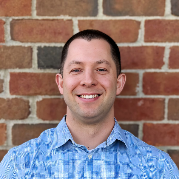Trent Wildman, Senior Living Coordinator at Holliday Farms Senior Living, smiling in a professional headshot, wearing a blue patterned button-down shirt, with a brick wall background.