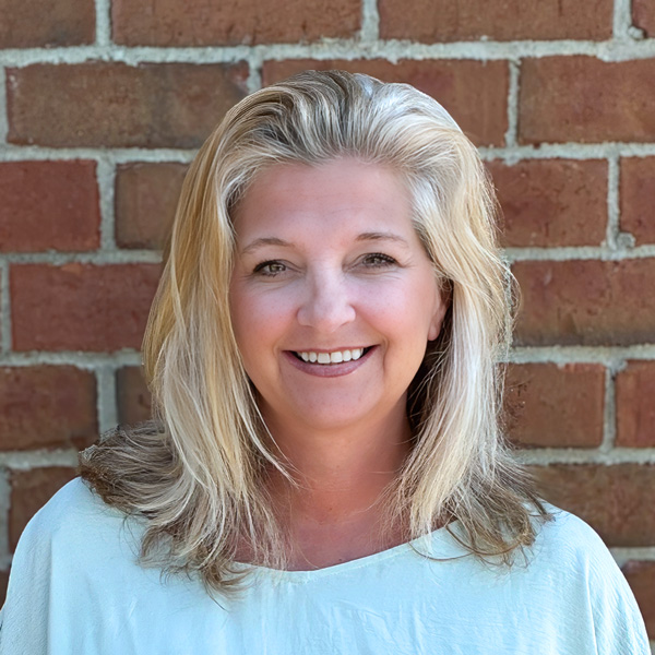 Brenda Irk, Resident Services Director at Holliday Farms Senior Living, smiling in a professional headshot with blonde hair, a light blouse, and a red brick wall background.
