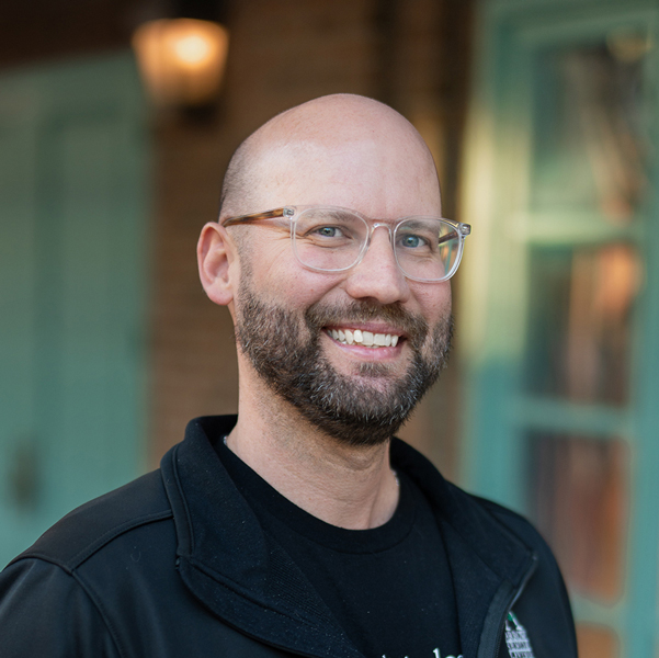 CJ Boswell, Executive Director at Holliday Farms Senior Living, smiling in a professional outdoor headshot with a bald head, clear glasses, a short beard, and softly blurred brick and teal window details in the background.