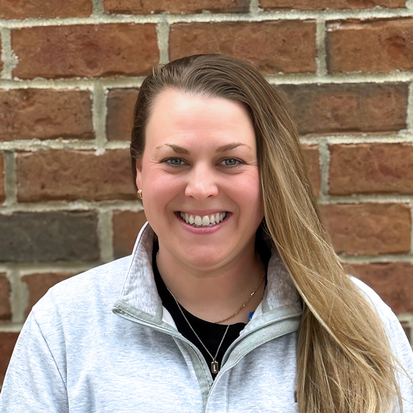 Lindsey Imel, Culinary Director at Holliday Farms Senior Living, smiling in a professional headshot with long light brown hair, a light quarter-zip pullover, and a red brick wall background.