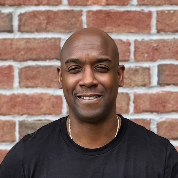 Jacobi Davis, Memory Care Director at Holliday Farms Senior Living, smiling in a professional headshot with a bald head, a black shirt, and a red brick wall background.