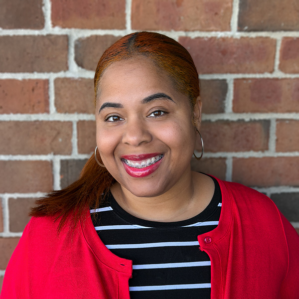 Ashlee Johnson, Wellness Director at Holliday Farms Senior Living, smiling in a professional headshot. She wears a red cardigan over a black-and-white striped top, with hoop earrings and a brick wall background.