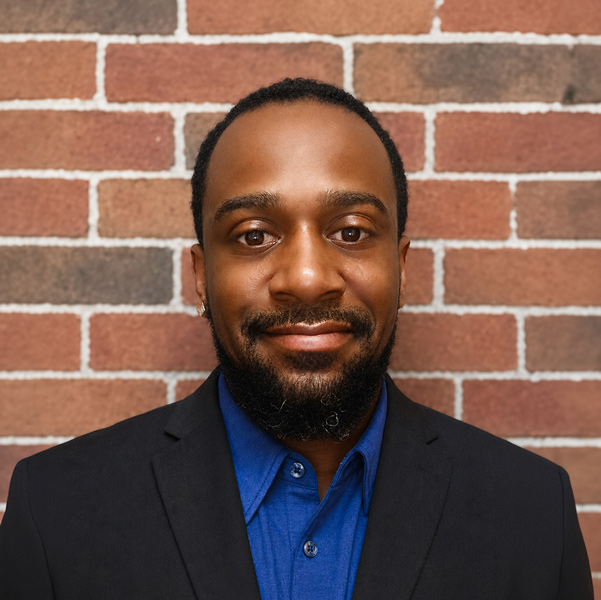 Daniel Thomas, Plant Operations Director at Holliday Farms Senior Living, looking at the camera in a professional headshot with short dark hair, a trimmed beard, a blue shirt, a black blazer, and a red brick wall background.