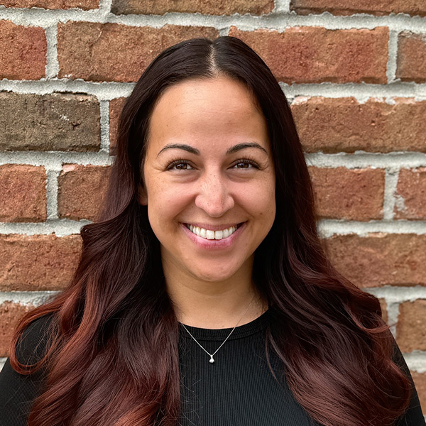 Jaynie Fukushima, Operations Coordinator at Holliday Farms Senior Living, smiling in a professional headshot with long dark auburn hair, a black top, a small pendant necklace, and a red brick wall background.