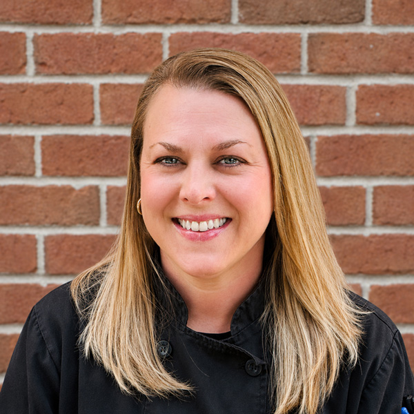 Lindsey Imel, Culinary Director at Holliday Farms Senior Living, smiling in a professional headshot wearing a black chef coat against a red brick wall background.