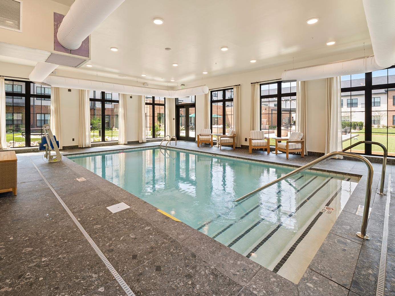 Bright indoor swimming pool at a senior living community with wide entry steps, handrails, an accessible lift, and poolside seating.