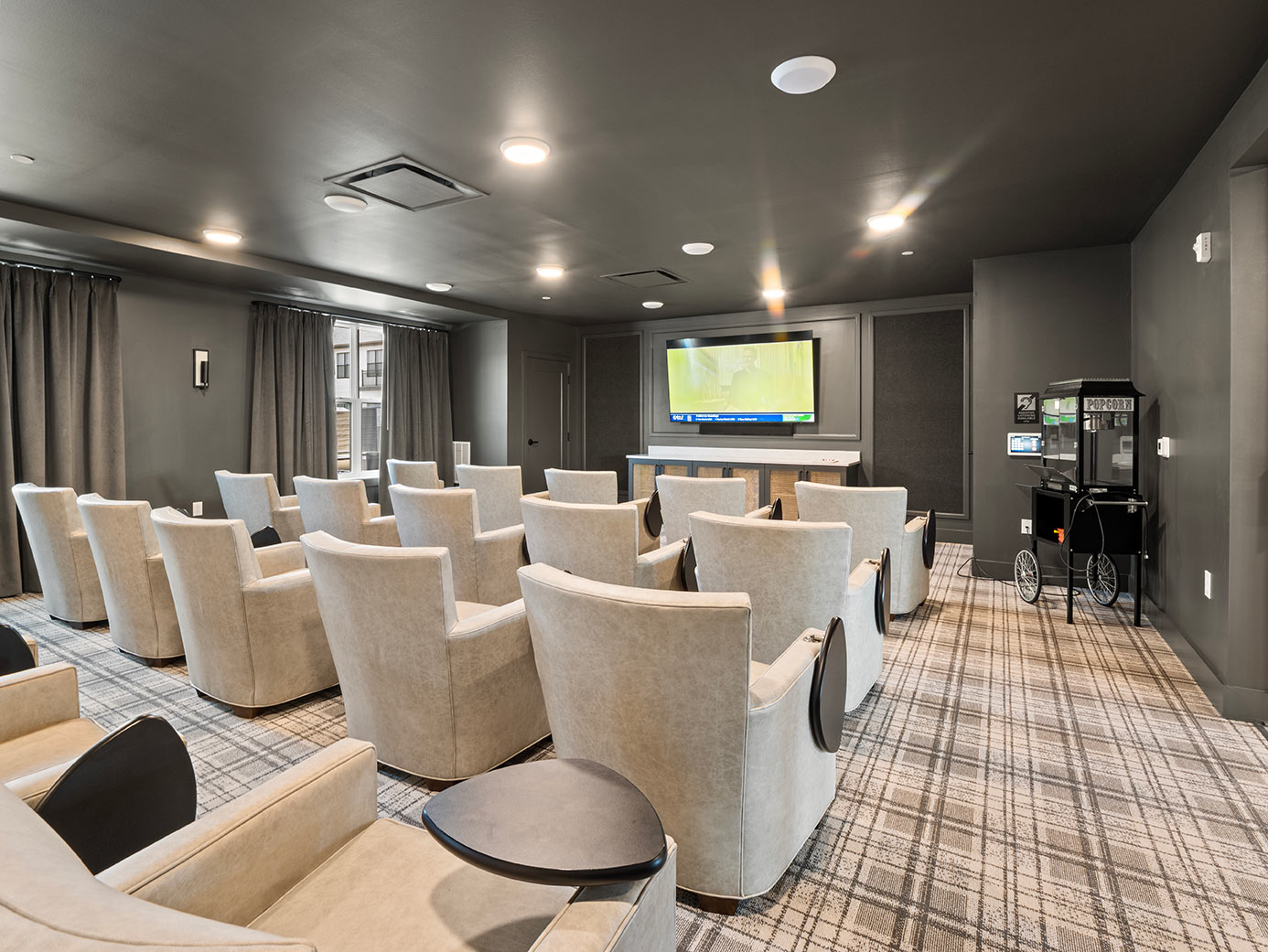 Senior living movie theater with comfortable upholstered seating, a large television, dark walls, and a popcorn machine.