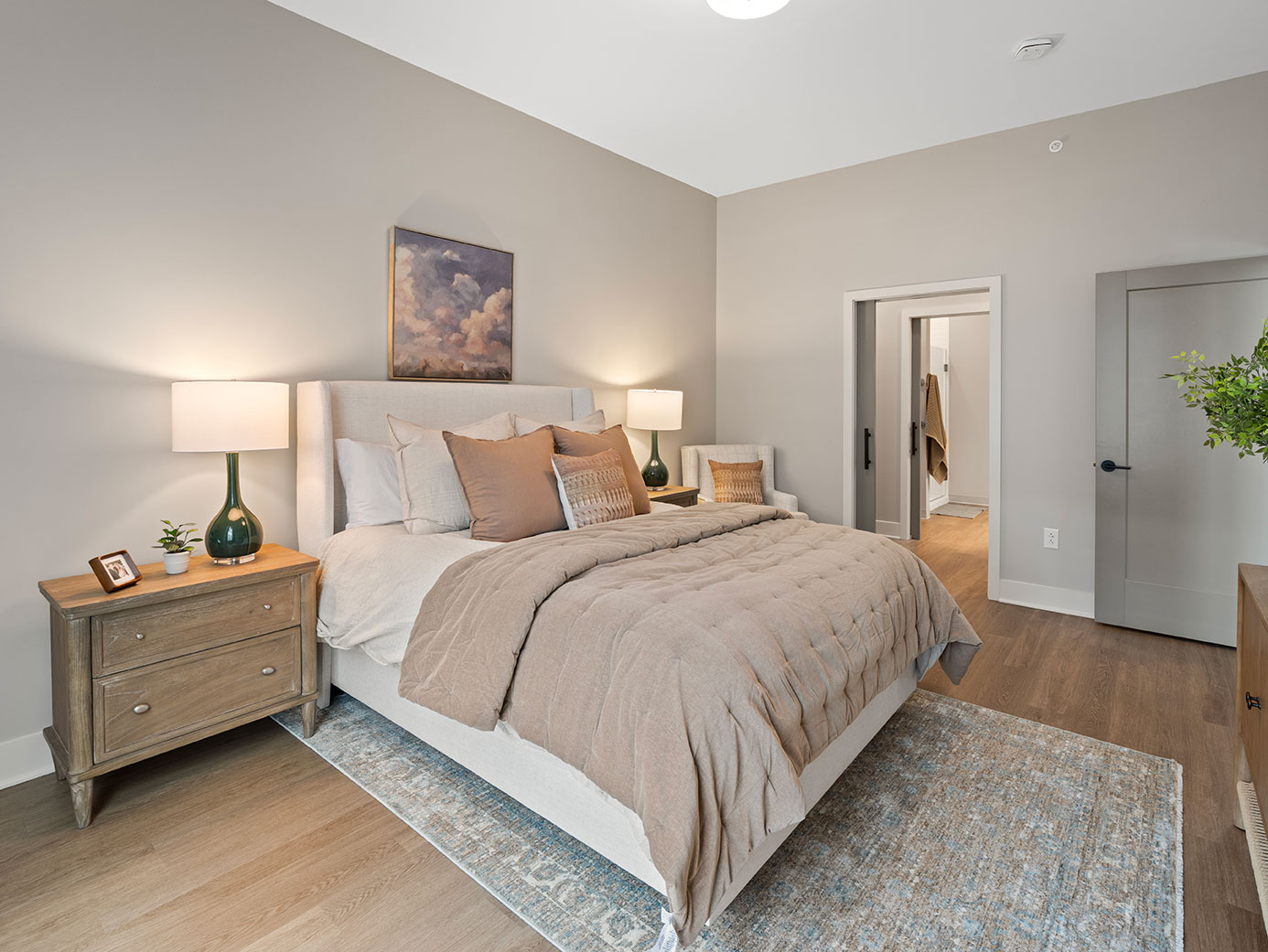 Spacious senior living apartment bedroom with a large upholstered bed, bedside tables, warm lighting, and an adjoining bathroom.