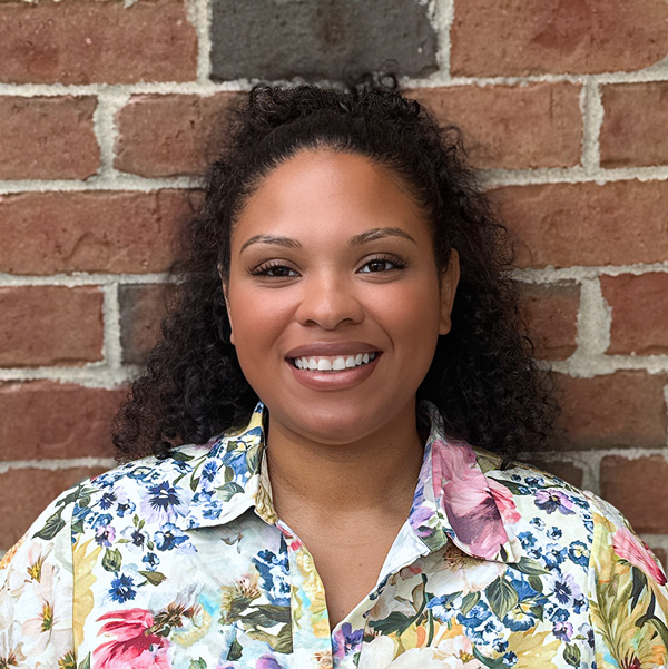 Johnea Goodman, Senior Living Coordinator at Holliday Farms Senior Living, smiling in a professional headshot with curly dark hair, a floral blouse, and a red brick wall background.